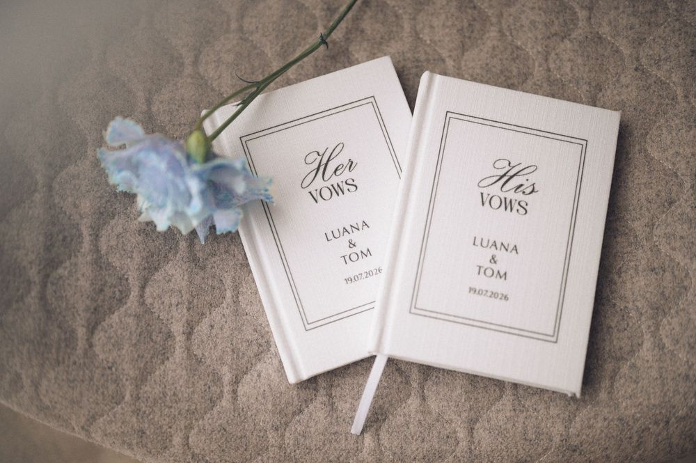 Personalized white wedding vow books for bride and groom, labeled His and Her Vows, displayed with a blue flower.
