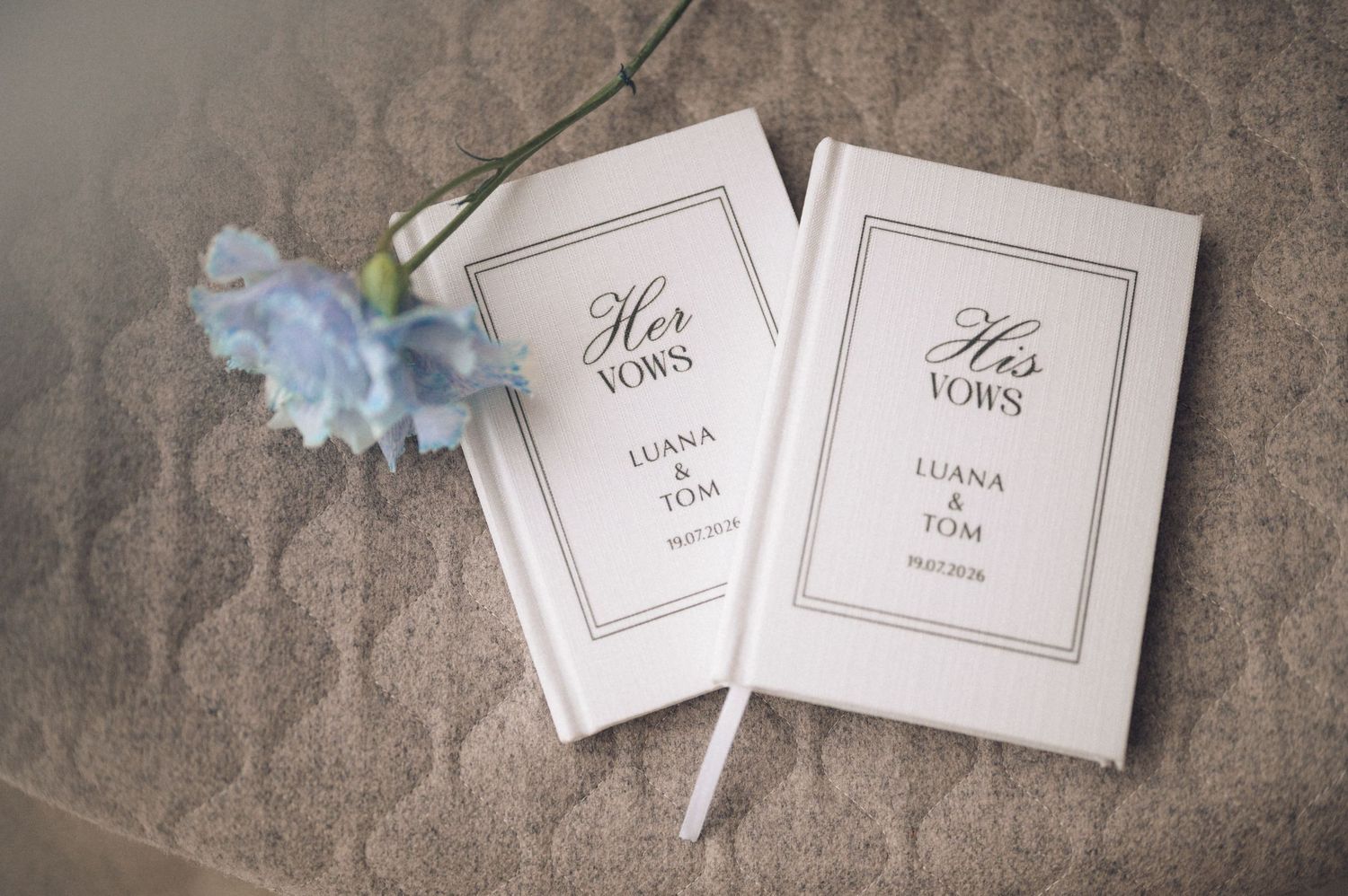 Personalized white wedding vow books for bride and groom, labeled His and Her Vows, displayed with a blue flower.