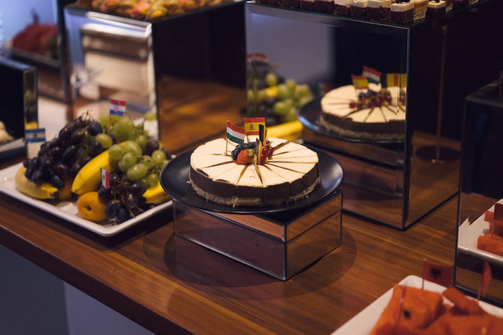 Elegant dessert display with a decorated cheesecake on a stand, fresh grapes, bananas, and watermelon on a wooden buffet table.