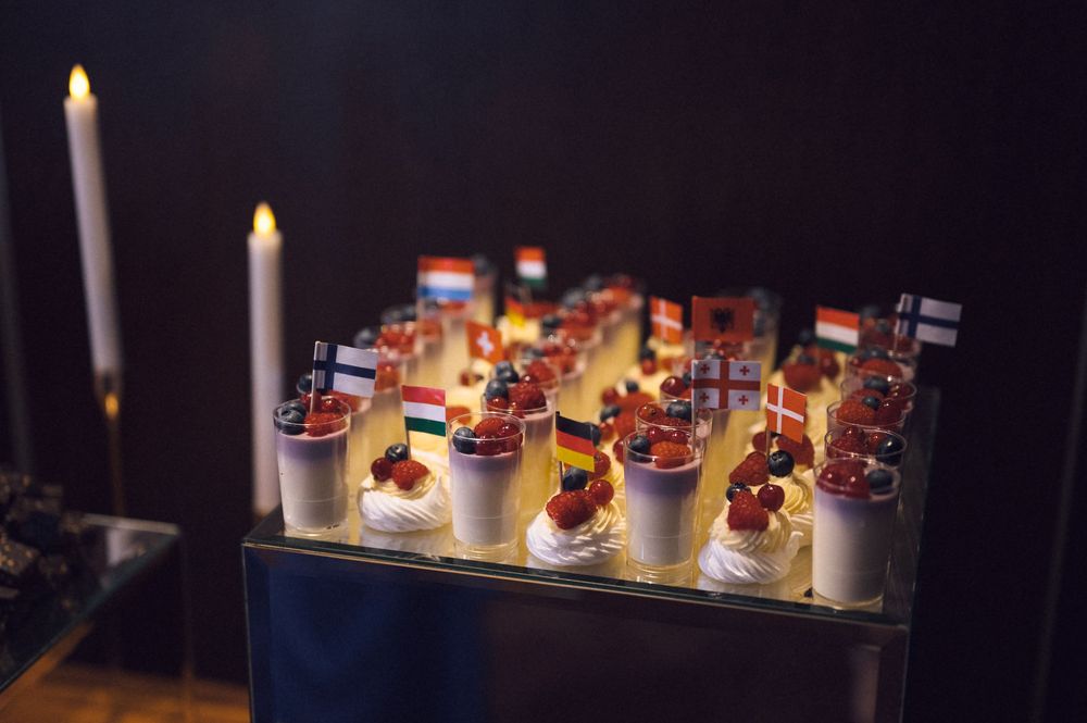 Elegant dessert cups topped with international flags displayed on a glass tray, flanked by candles at a formal event.