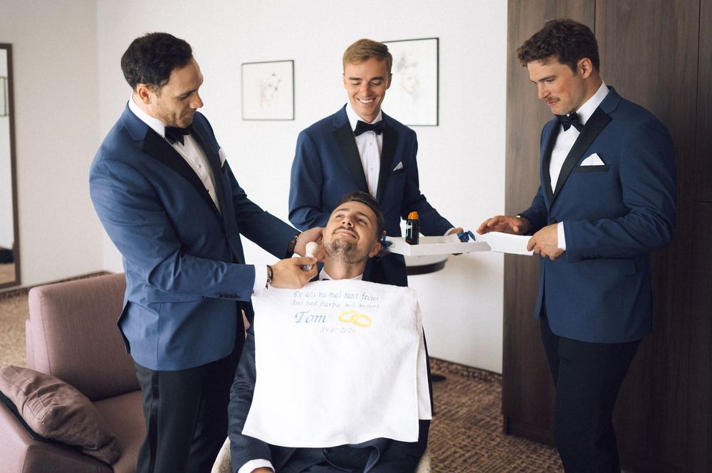 Groomsmen in navy tuxedos help groom put on shirt before wedding ceremony indoors.