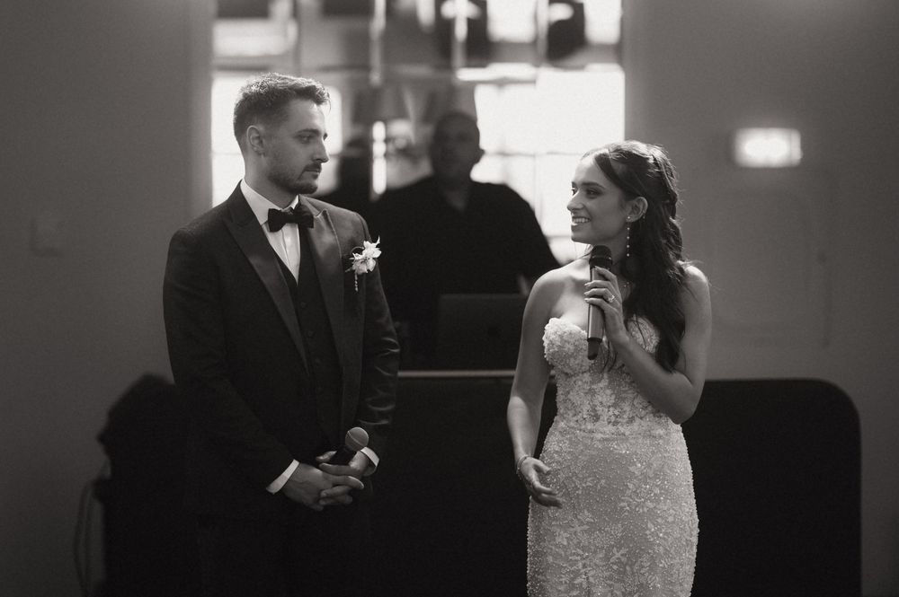 Black and white photo of bride speaking at microphone beside groom in tuxedo during wedding reception.