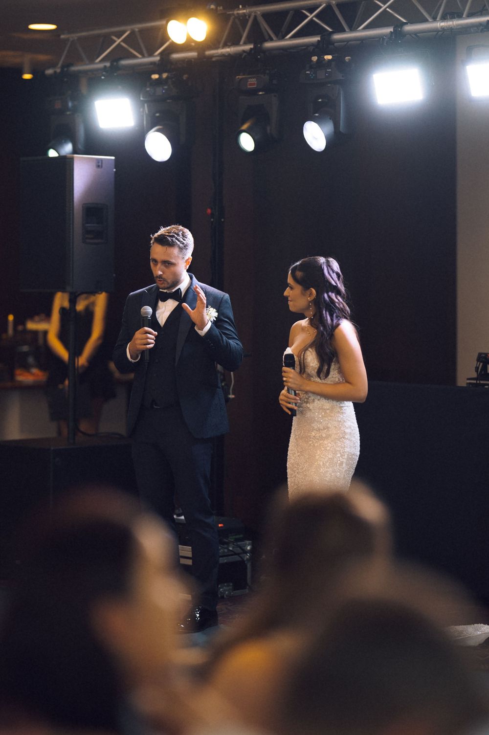 Bride and groom sharing microphones on stage during wedding reception speech under stage lighting.