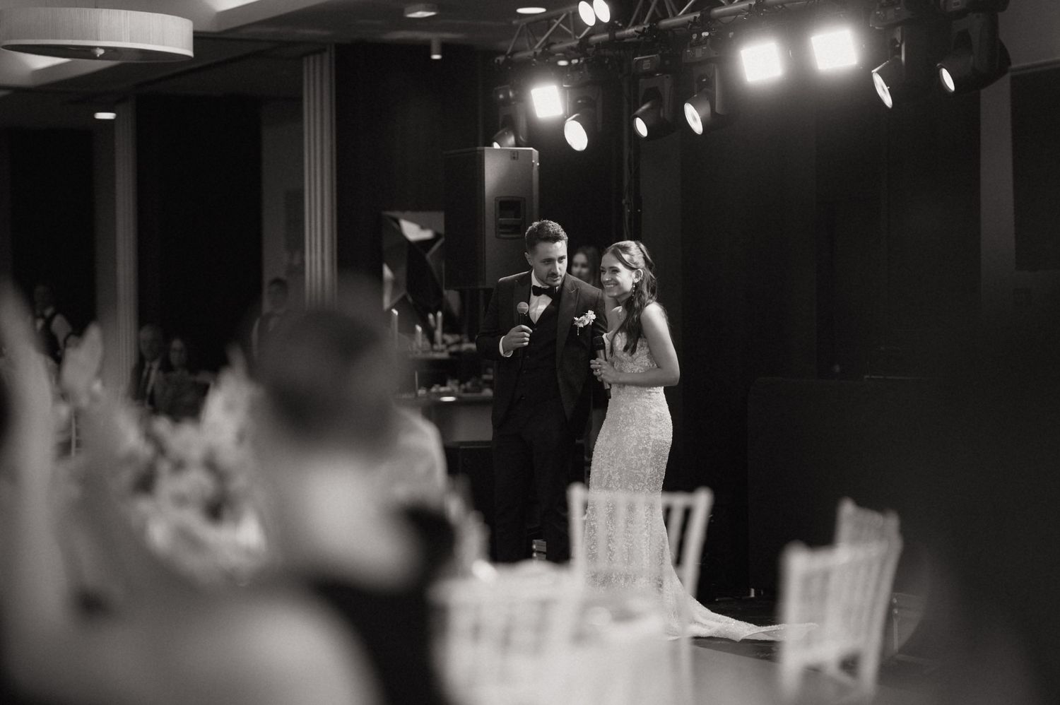 Black and white photo of bride and groom sharing first dance at elegant wedding reception with stage lighting.