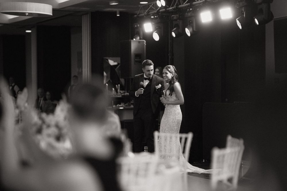 Black and white photo of bride and groom sharing first dance at elegant wedding reception with stage lighting.