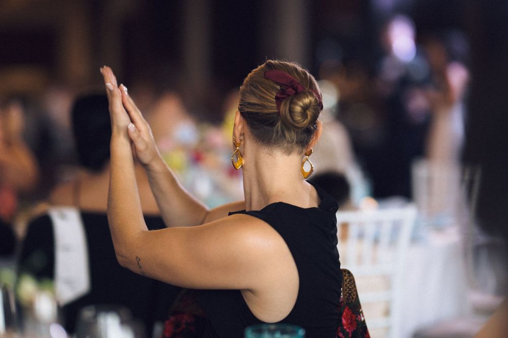 Woman in black dress with floral detail clapping at an elegant indoor event, hair in a bun with red accessory.