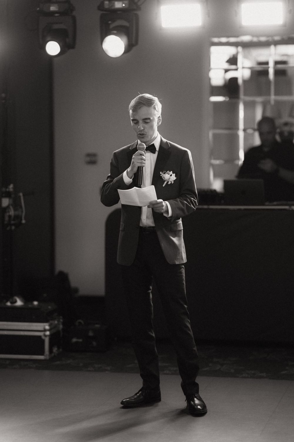 Young man in suit reading speech at wedding reception, holding microphone and paper, black and white photo.