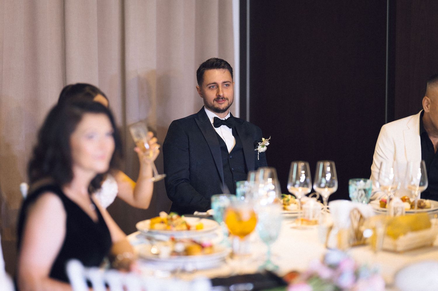 Groom in black tuxedo and bow tie listens attentively during wedding reception dinner toast.