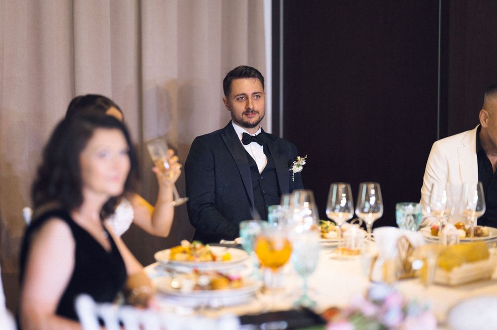 Groom in black tuxedo and bow tie listens attentively during wedding reception dinner toast.