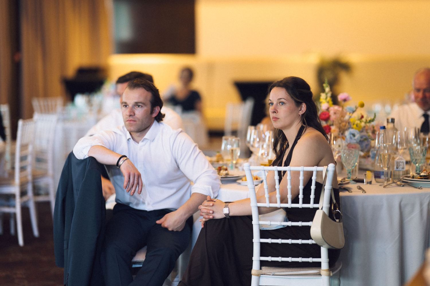 Man and woman sitting at elegantly decorated wedding reception table, looking thoughtfully into the distance.