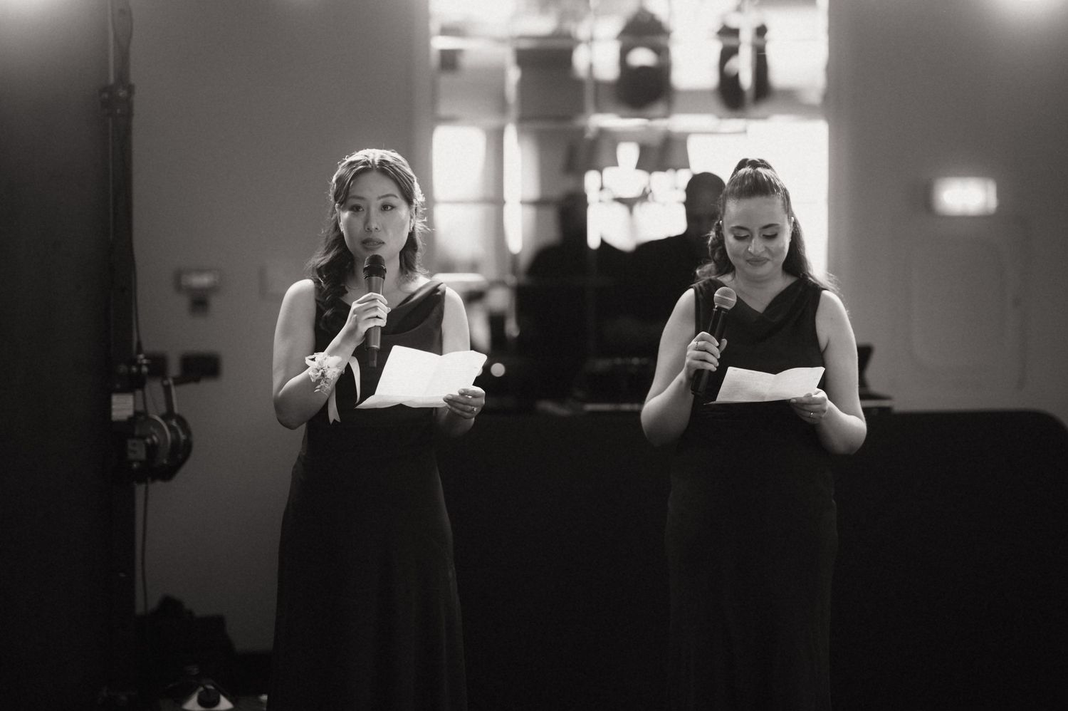 Two women in black dresses reading from papers while speaking into microphones at a formal event.