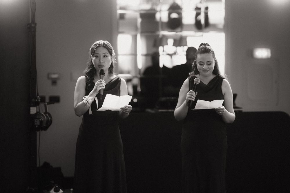 Two women in black dresses reading from papers while speaking into microphones at a formal event.