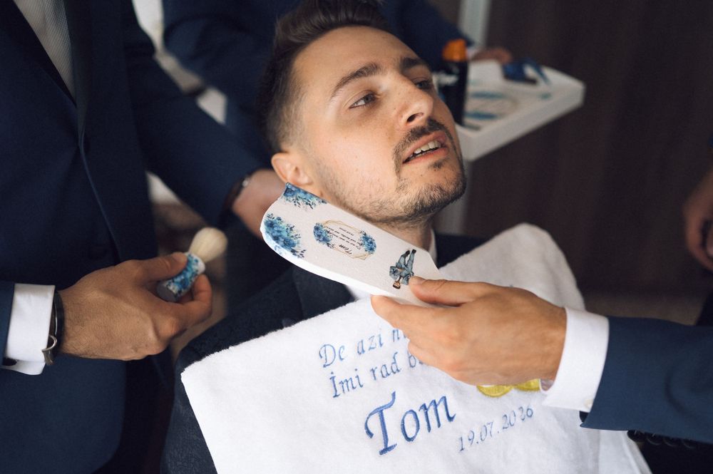 Man receiving a shave at a barbershop, draped with an embroidered cloth reading 'Tom'.