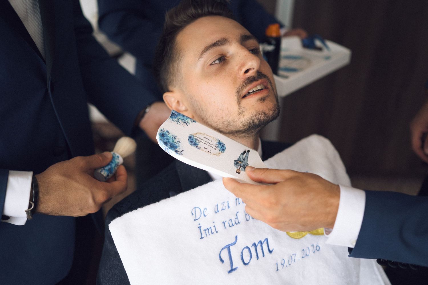 Man receiving a shave at a barbershop, draped with an embroidered cloth reading 'Tom'.
