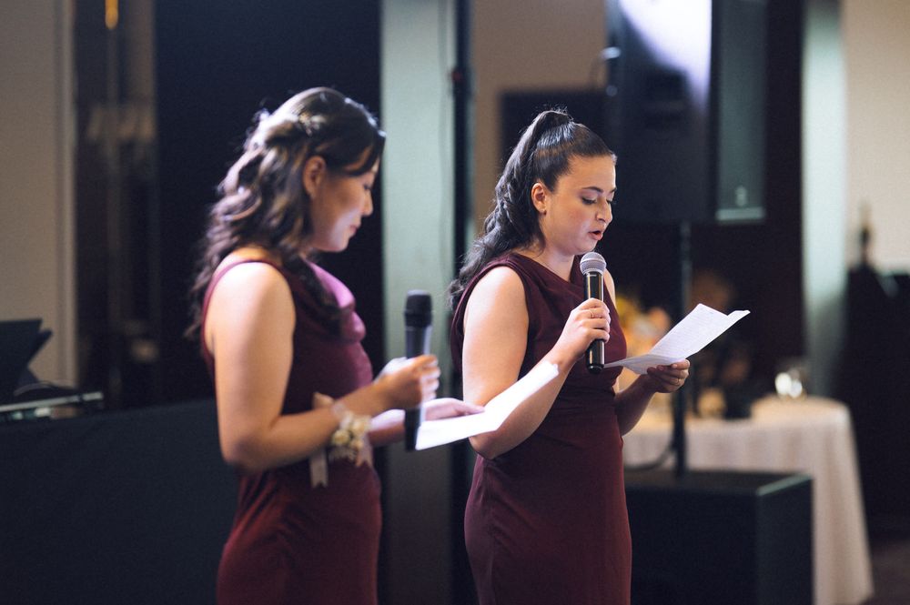 Two women in burgundy dresses giving a speech at a wedding reception, one reading from paper while holding a microphone.