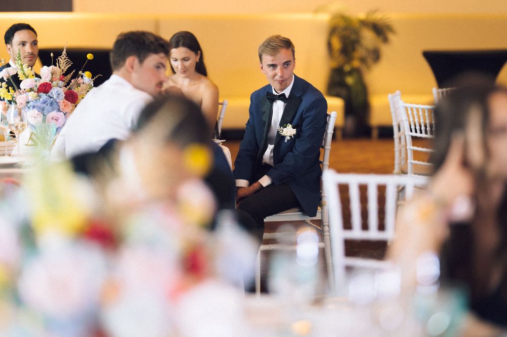 Groom in navy suit sits attentively at wedding reception table surrounded by guests and colorful floral centerpieces.
