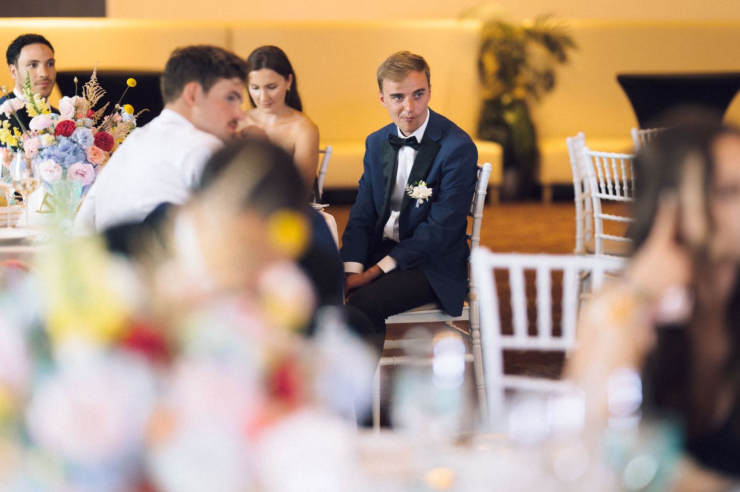 Groom in navy suit sits attentively at wedding reception table surrounded by guests and colorful floral centerpieces.
