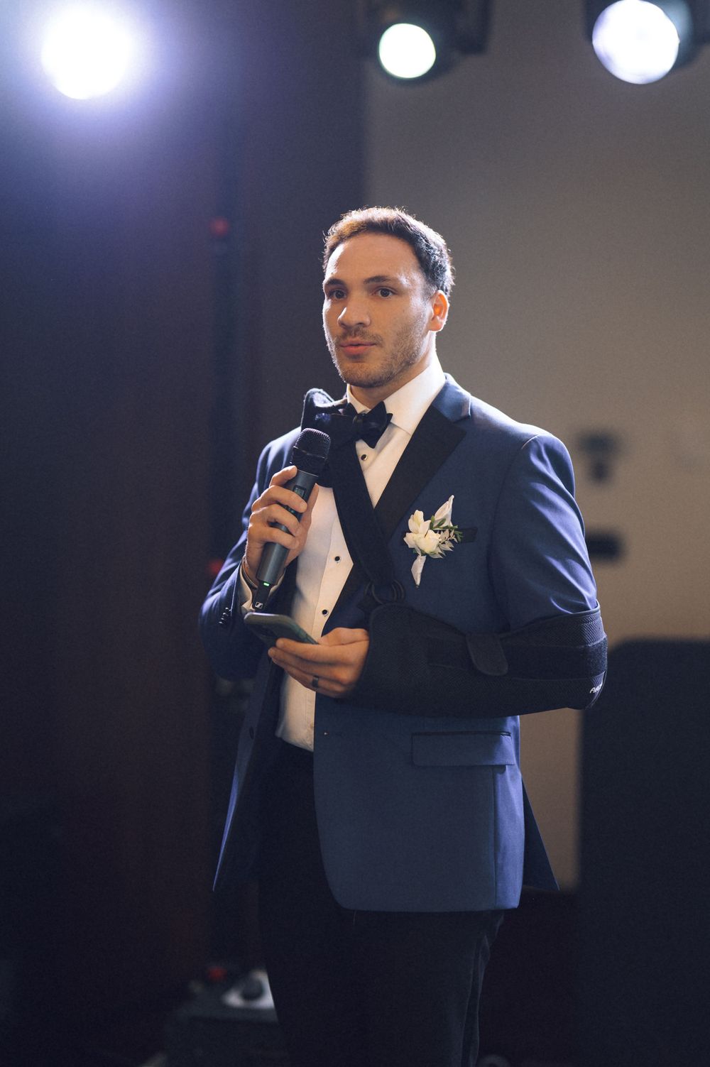 Man in navy tuxedo with boutonniere speaks into microphone at formal event under stage lighting.