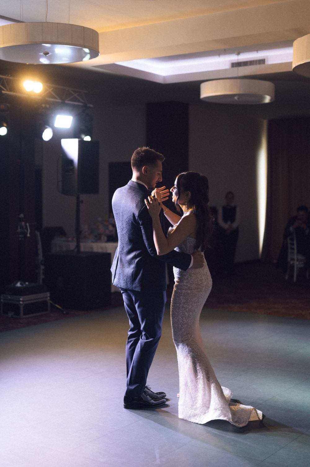 Bride and groom share their first dance in an elegant ballroom, dressed in a suit and fitted white gown.
