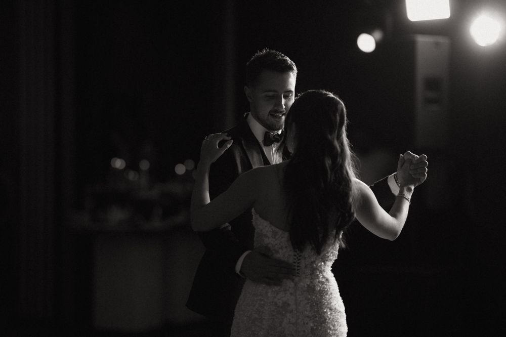 Black and white photo of a couple sharing their first dance at a wedding reception, illuminated by soft stage lighting.