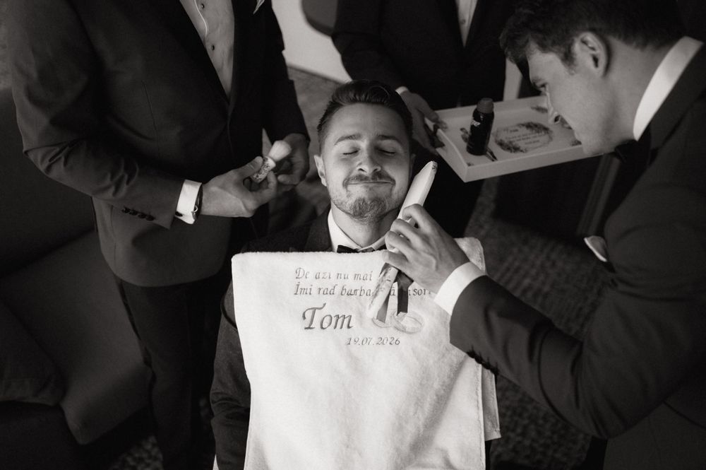Groom relaxes in chair as groomsmen assist with wedding preparations in black and white photo.