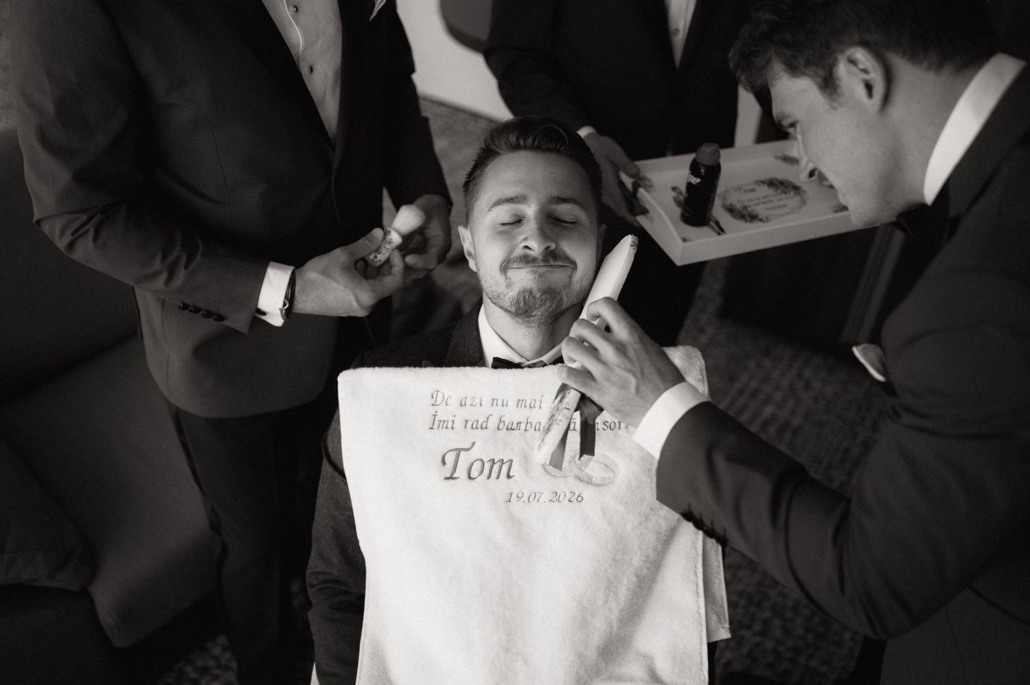 Groom relaxes in chair as groomsmen assist with wedding preparations in black and white photo.
