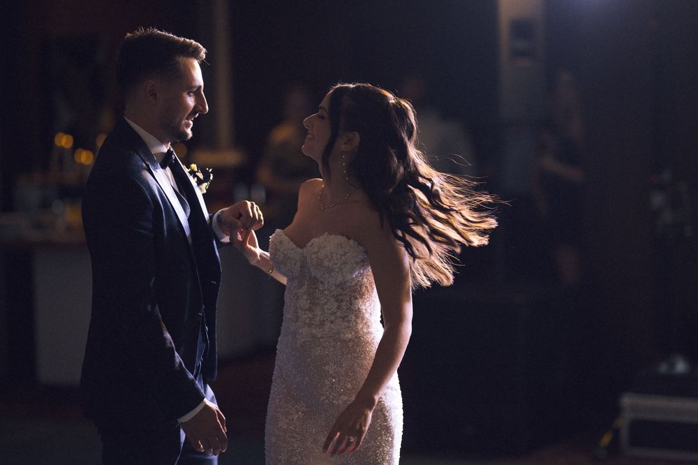 Bride and groom share romantic first dance, bride in lace gown, groom in dark suit, soft dramatic lighting.