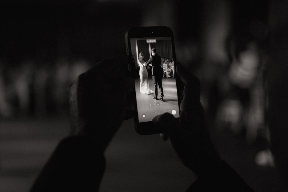 Black and white photo of hands holding a smartphone capturing a couple's first dance at a wedding.