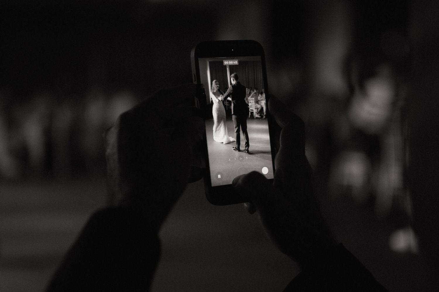 Black and white photo of hands holding a smartphone capturing a couple's first dance at a wedding.