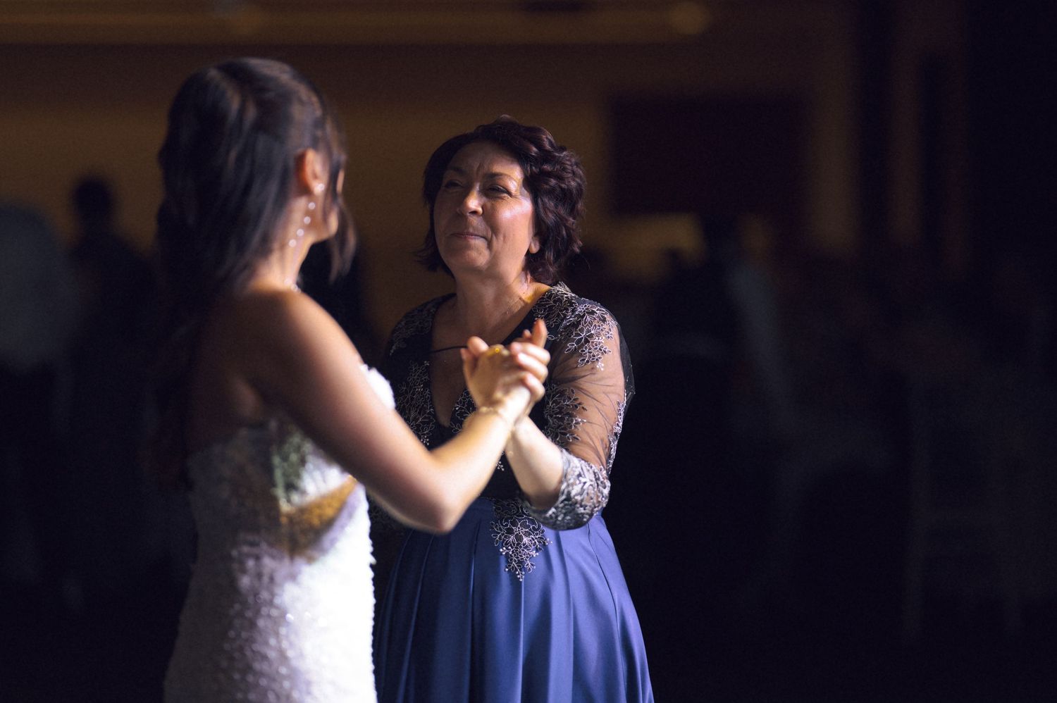 Two women share an emotional dance at a wedding reception, one in a white bridal gown and the other in a blue dress.