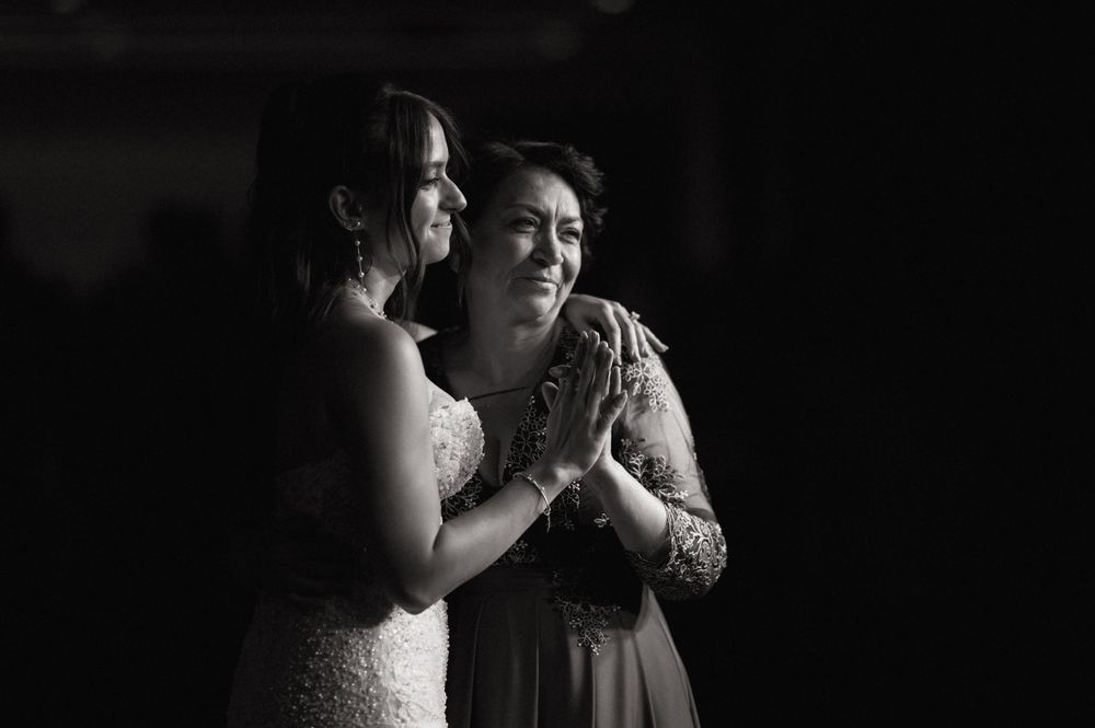 Black and white photo of bride and older woman sharing emotional moment, hands clasped together at wedding reception.