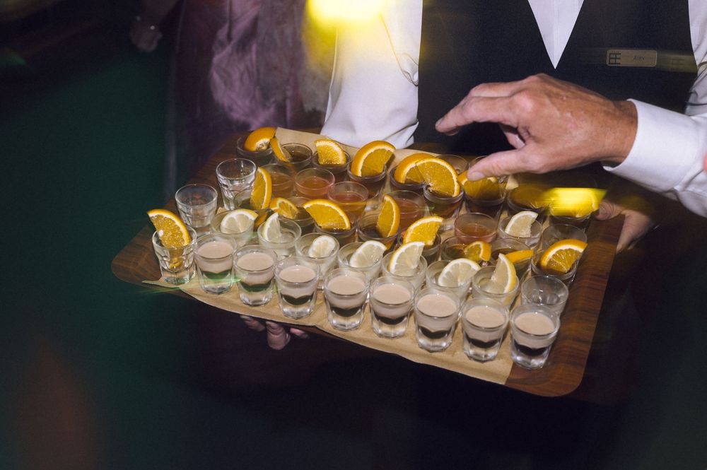 Waiter holding a wooden tray of shot glasses garnished with orange and lemon slices at a catered event.