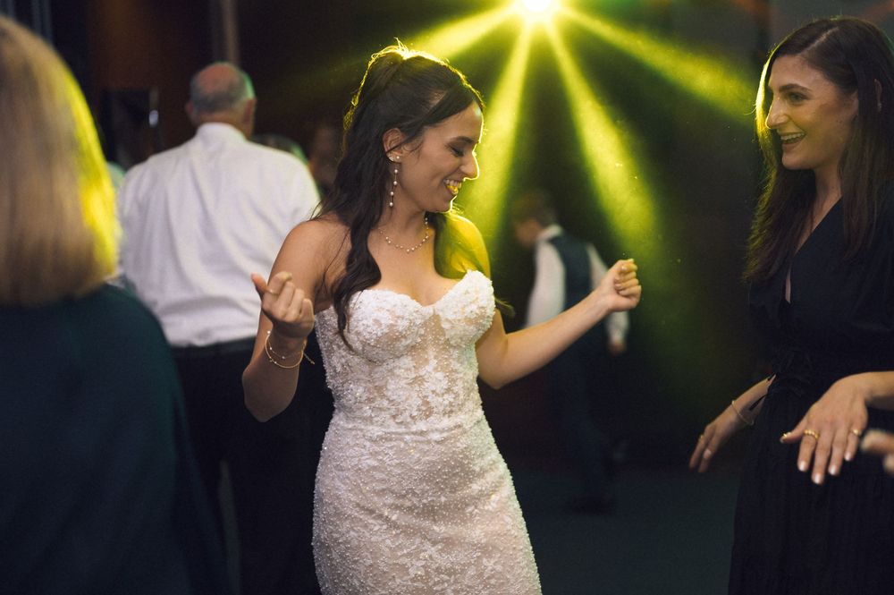 Bride in white lace gown dancing joyfully with guests at a wedding reception under green stage lights.
