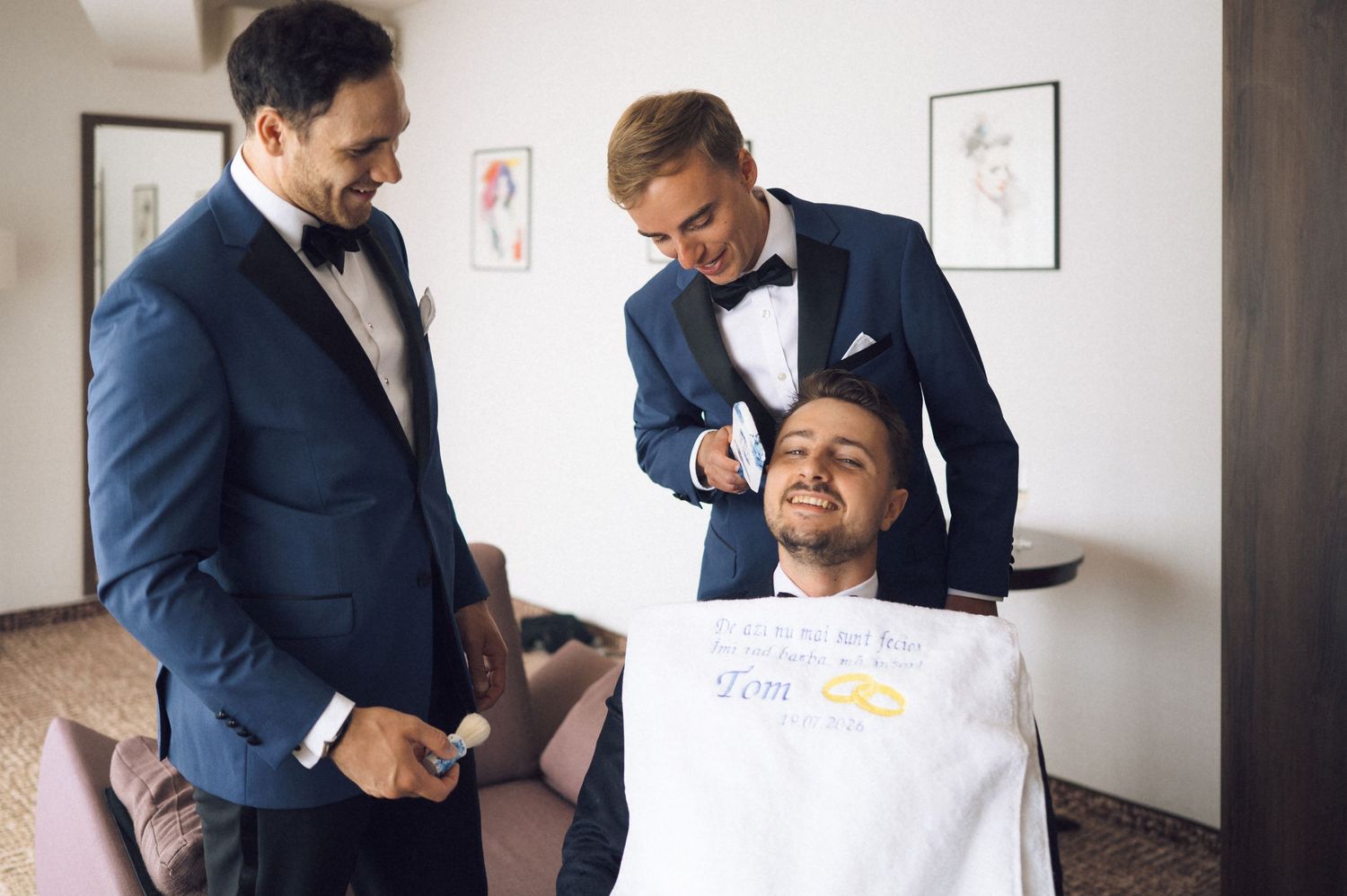 Two groomsmen in navy tuxedos help a seated groom prepare for his wedding day indoors.