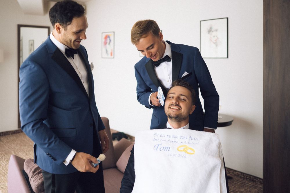 Two groomsmen in navy tuxedos help a seated groom prepare for his wedding day indoors.