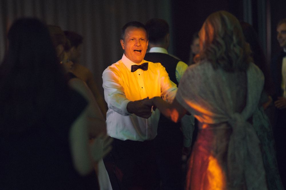Man in tuxedo dancing joyfully at formal event with guests in elegant attire under warm lighting.