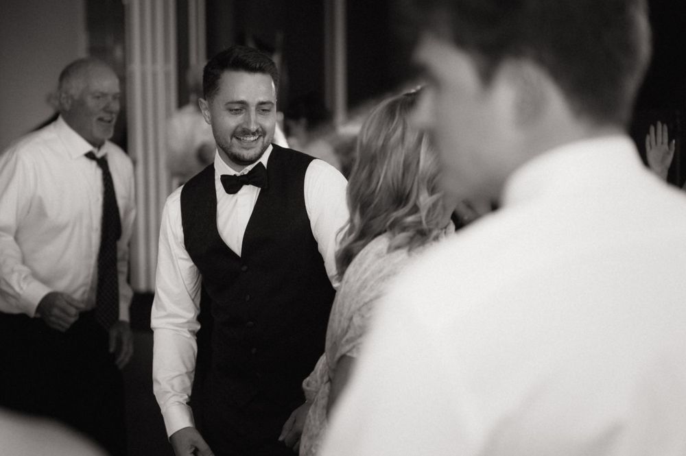 Black and white photo of a smiling groom in bow tie and vest dancing at a wedding reception.