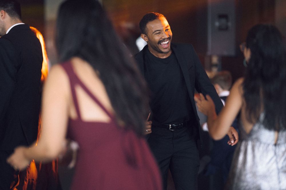 A man in black laughs joyfully while dancing at a formal event surrounded by elegantly dressed guests.