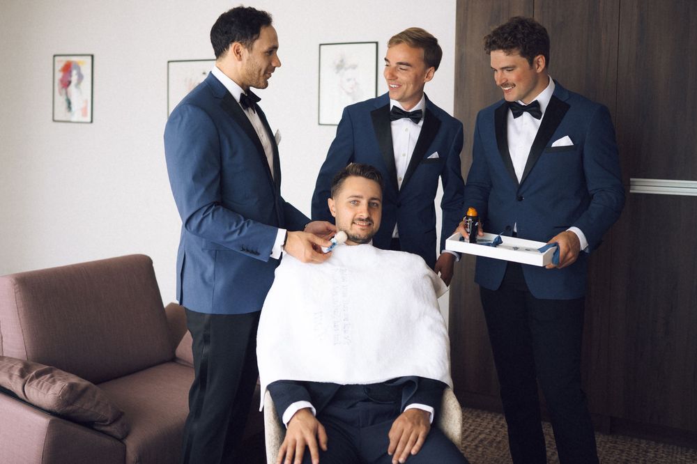 Four men in navy tuxedos share a lighthearted grooming moment before a formal event.