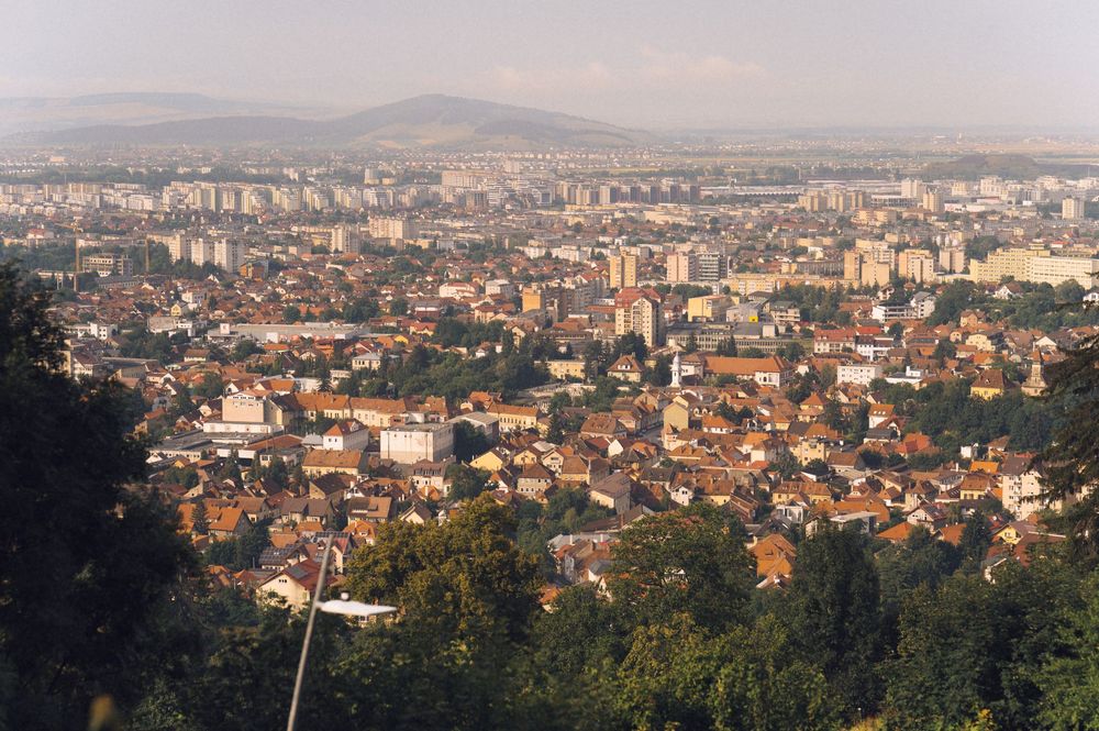 Aerial panoramic view of a European city with red-roofed buildings, modern tower blocks, and green hills in the background.