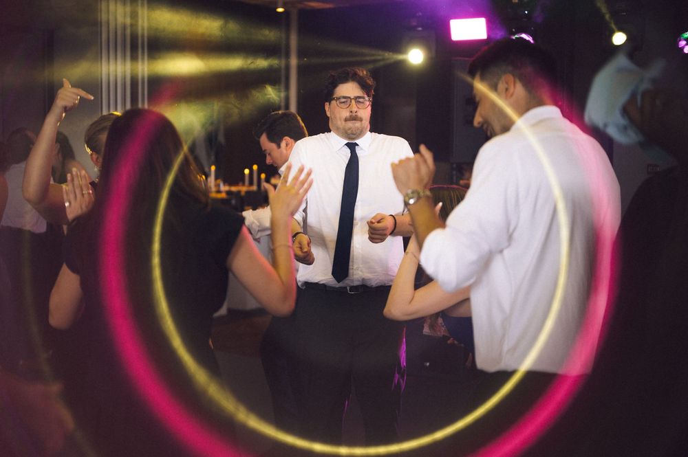 Man in white shirt and tie dancing at a lively party with colorful light trails and guests surrounding him.