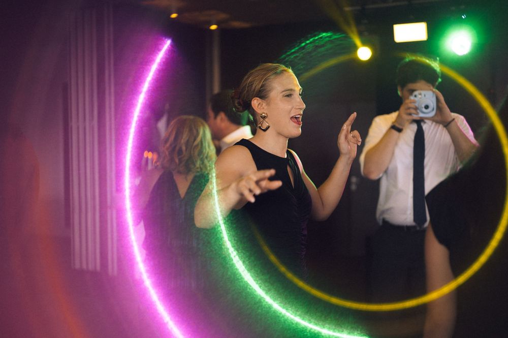 Woman dancing at a party surrounded by colorful neon hoop lights, with a photographer capturing the moment in the background.