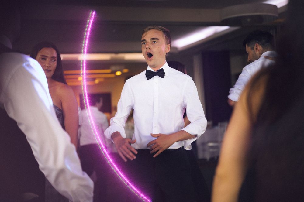 Young man in white shirt and bow tie dancing at a formal event with pink neon light in background.
