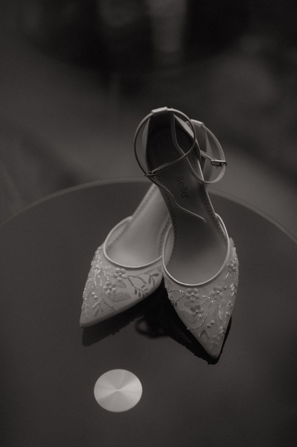 Black and white photo of elegant lace bridal heels with ankle straps displayed on a reflective table.