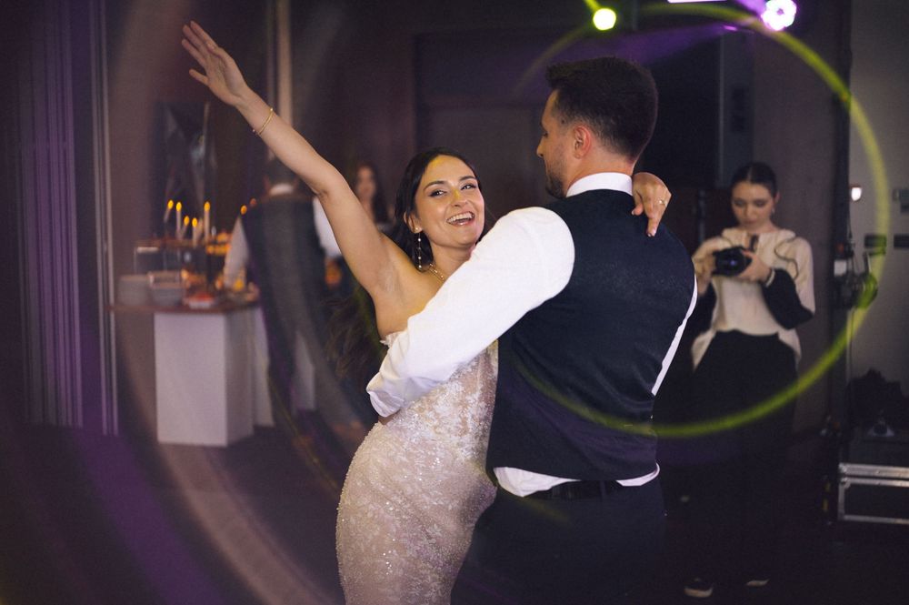 Bride and groom share their first dance at a wedding reception, with guests capturing the moment in the background.