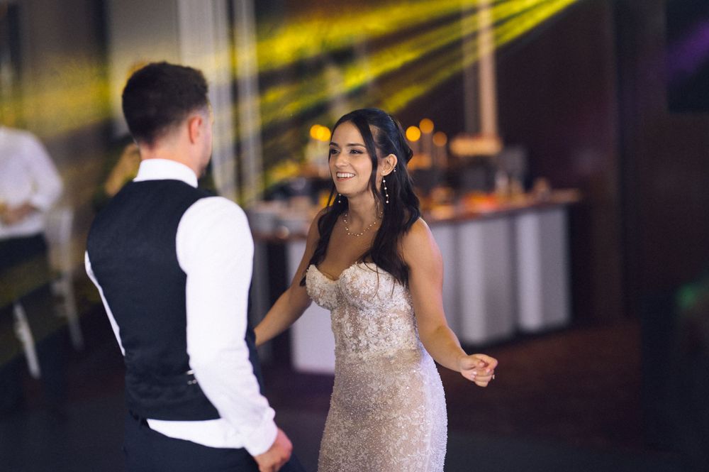 Bride in beaded gown shares first dance with groom in black vest under colorful lights at wedding reception.