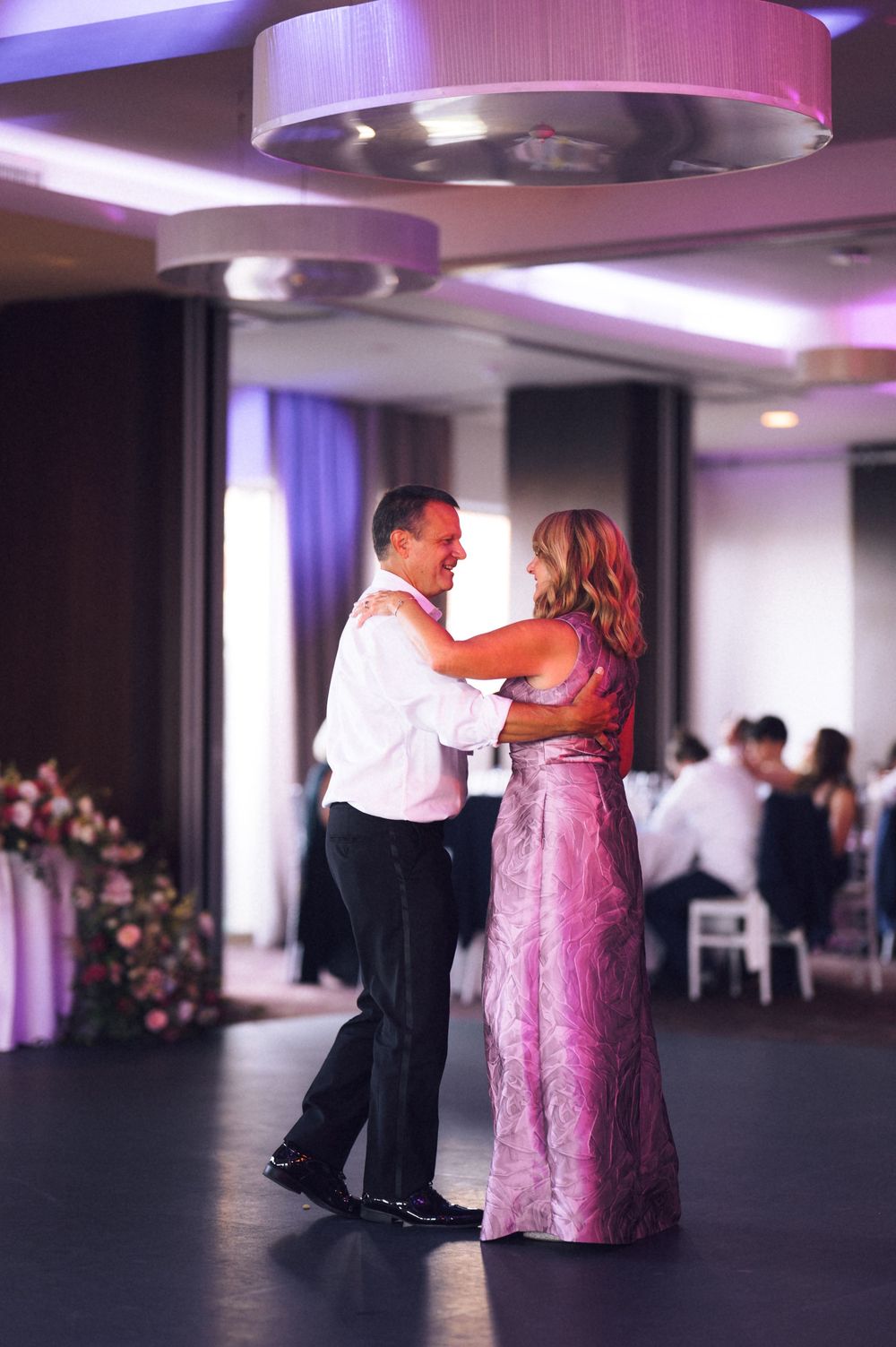 Couple dancing elegantly at a wedding reception in a ballroom with purple lighting, man in white shirt, woman in pink gown.