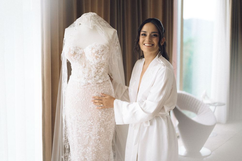 Bride in white robe smiling beside elegant lace wedding dress with veil on mannequin in bright room.