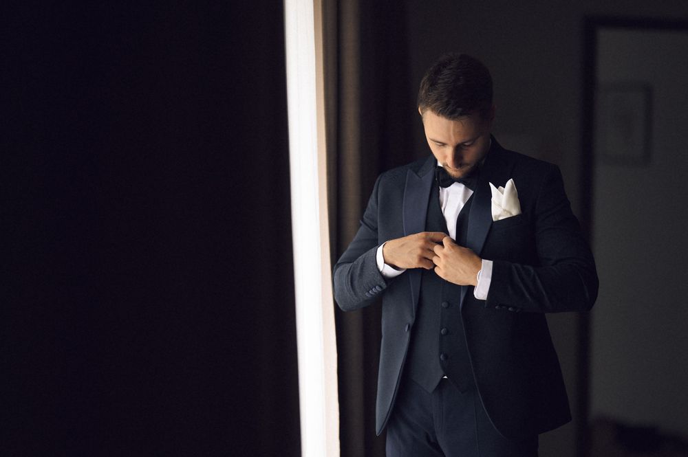 Elegant groom in a dark navy suit adjusting his bow tie while standing near a bright window.