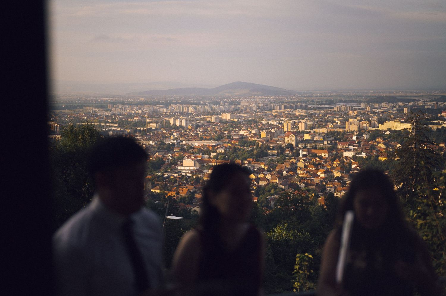 Blurred figures stand on a hilltop overlook with a panoramic cityscape and mountain backdrop at dusk.